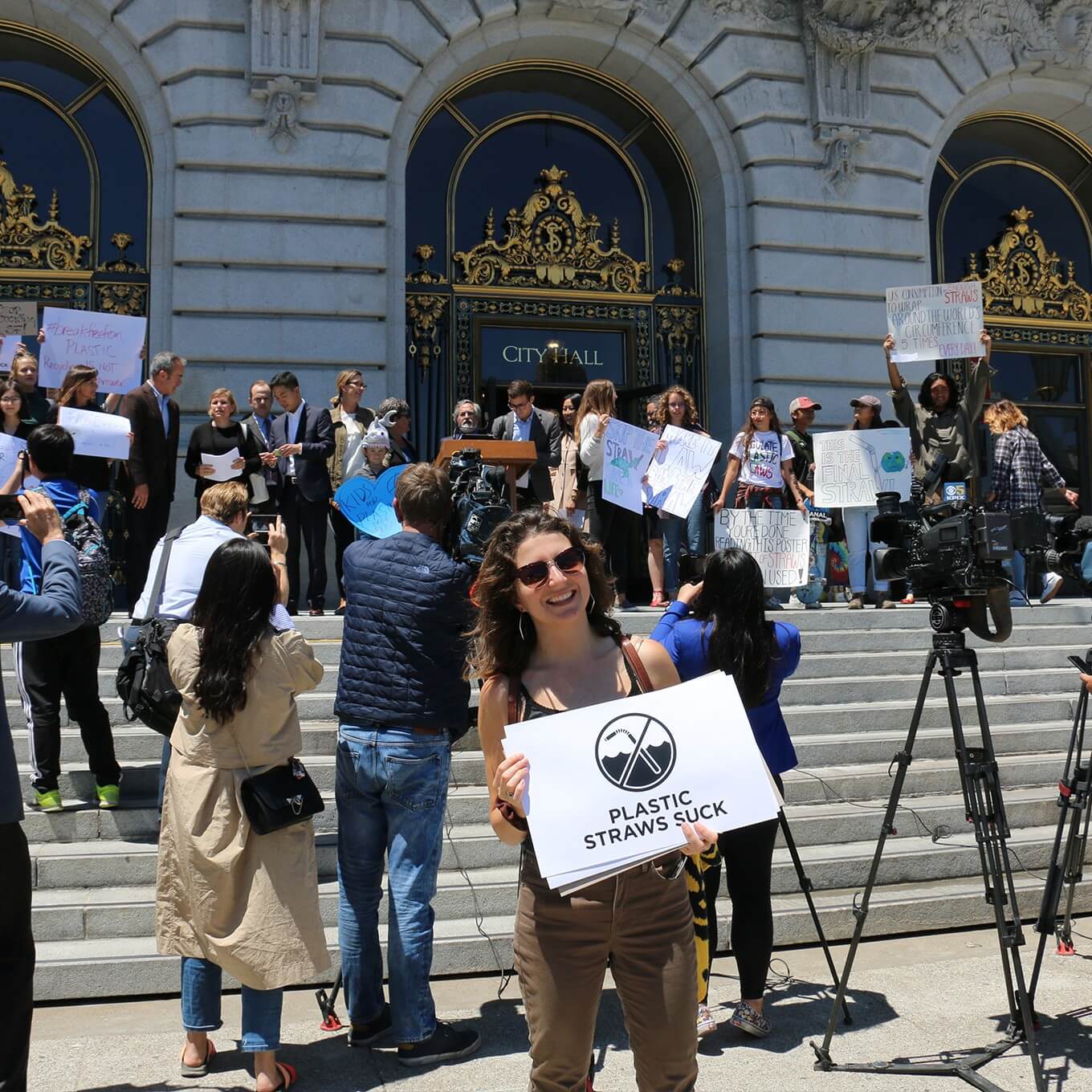 woman in front of city hall building with a sign that reads "plastic straws suck"
